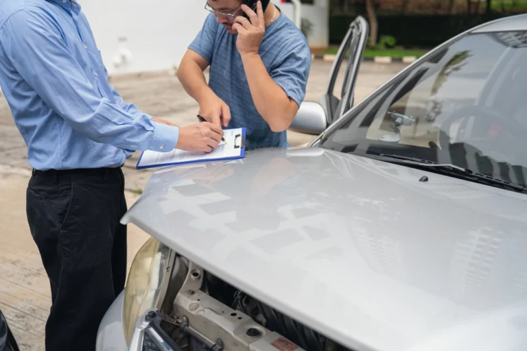 Two men inspecting a damaged vehicle with open hood, one on the phone and the other taking notes, illustrating a Florida car accident claim settlement and insurance evaluation process.